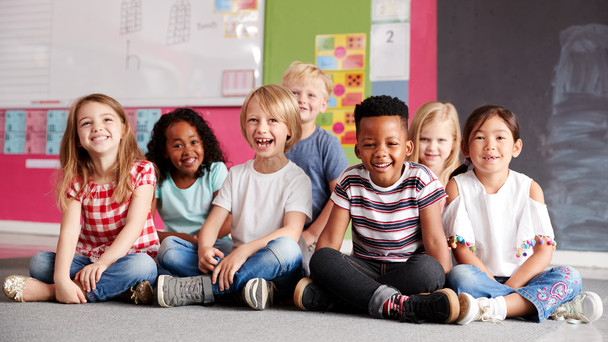 Lachende Schülerinnen und Schüler sitzen auf dem Fußboden eines Klassenzimmers.